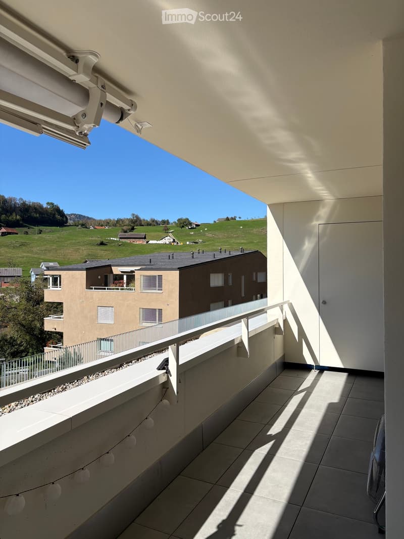 Balcony with tiled floor, white railing, and a view of a green landscape and neighboring buildings.