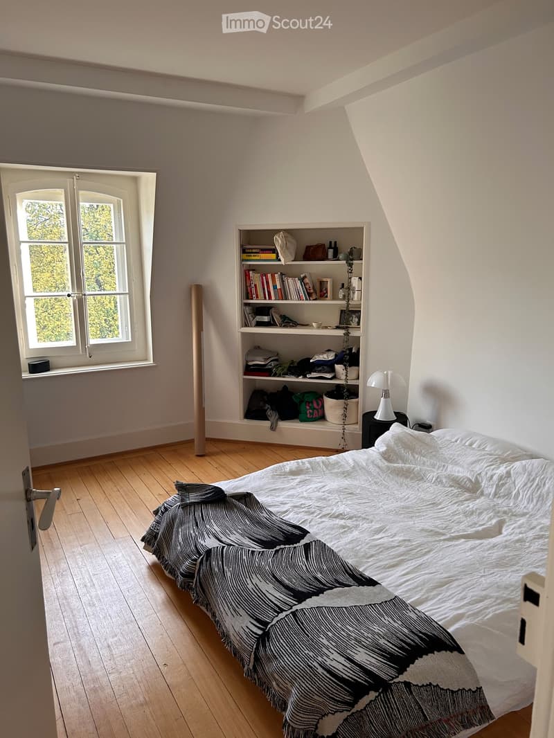Cozy attic bedroom with wooden floors, large window, bed with patterned blanket, bookshelf, and small table with lamp.