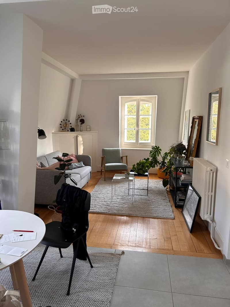 Bright living room with wooden flooring, large window, grey sofa, armchair, coffee table, and several plants. Includes a small dining area with a round table and chair.
