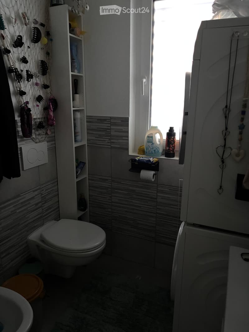 A small bathroom with a toilet, sink, and shower. A shelf holds various toiletries, and a window lets in natural light. Toilet paper is on a holder near the window, and a bottle is placed on the windowsill.