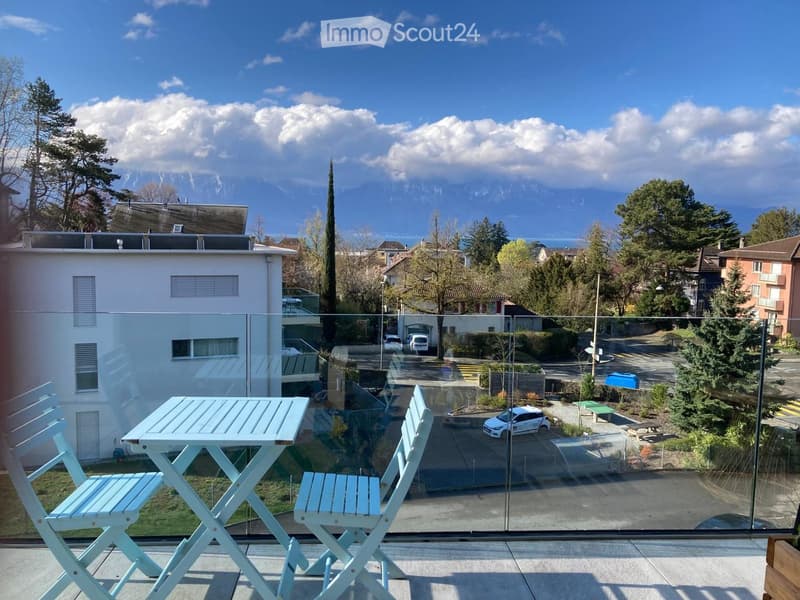 Balcony with a glass railing, a small table, and two chairs, overlooking a scenic view of mountains and a town.