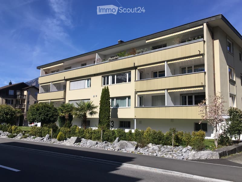 A modern multi-story apartment building with balconies, large windows, and a landscaped garden area in front.