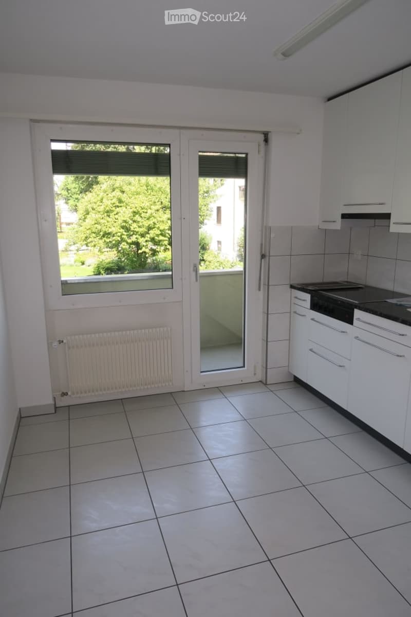 Empty kitchen with white tiled floor, white cabinets, and a large window with a view of greenery outside.