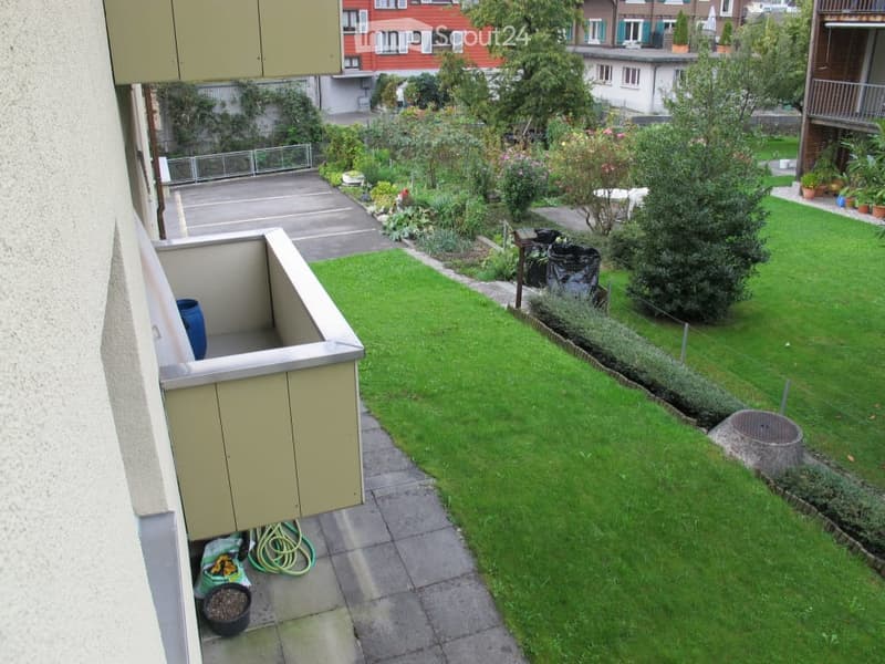 Balcony with small seating area, green lawn, garden beds, and surrounding buildings visible.