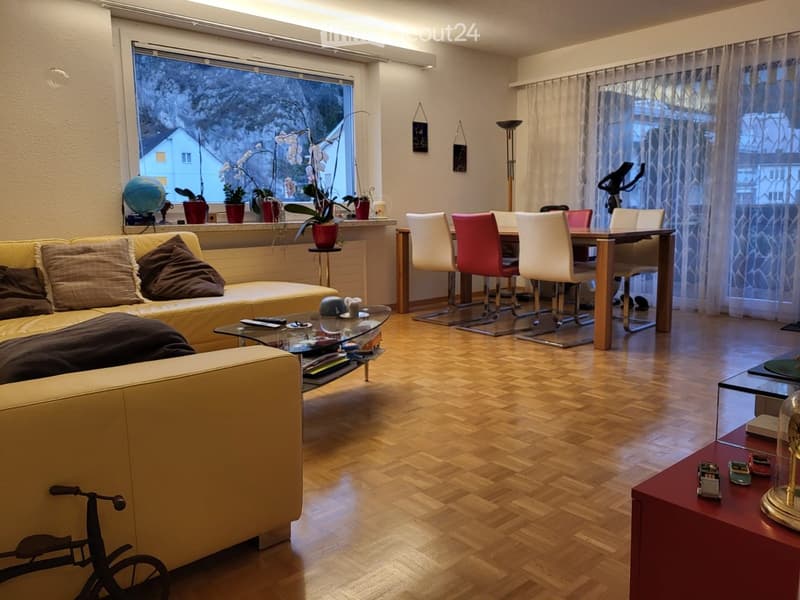 Living room with a cream-colored sofa, glass coffee table, dining table with chairs, potted plants, and a bicycle in the corner.