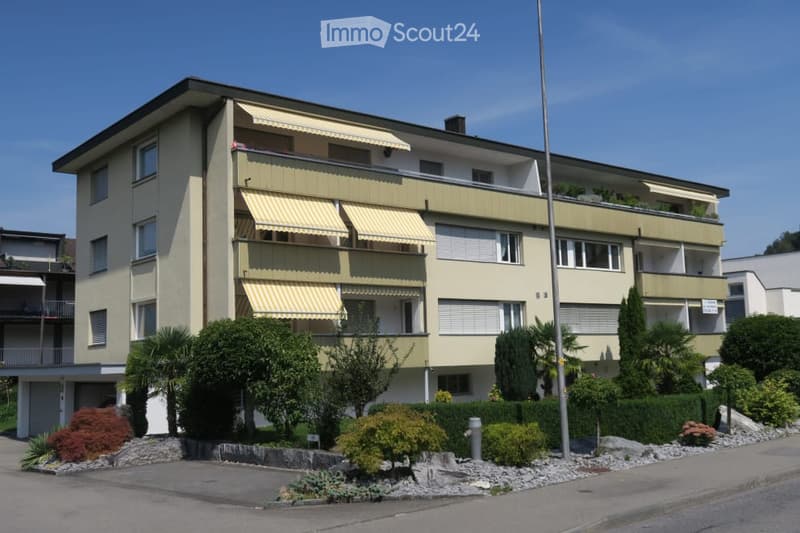 A multi-story residential building with beige walls, multiple windows, and balconies with yellow awnings. The building is surrounded by well-maintained landscaping with trees and shrubs.