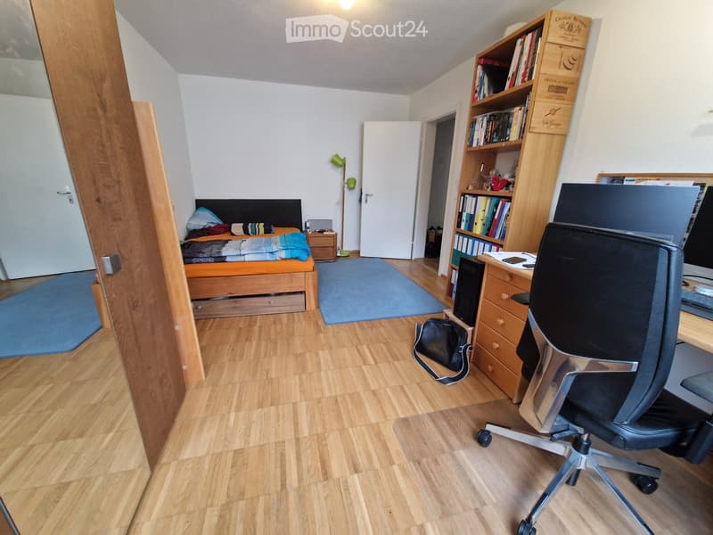 A furnished room with a bed, desk, chair, bookshelf, and a lamp. The room features wooden flooring and a blue rug.