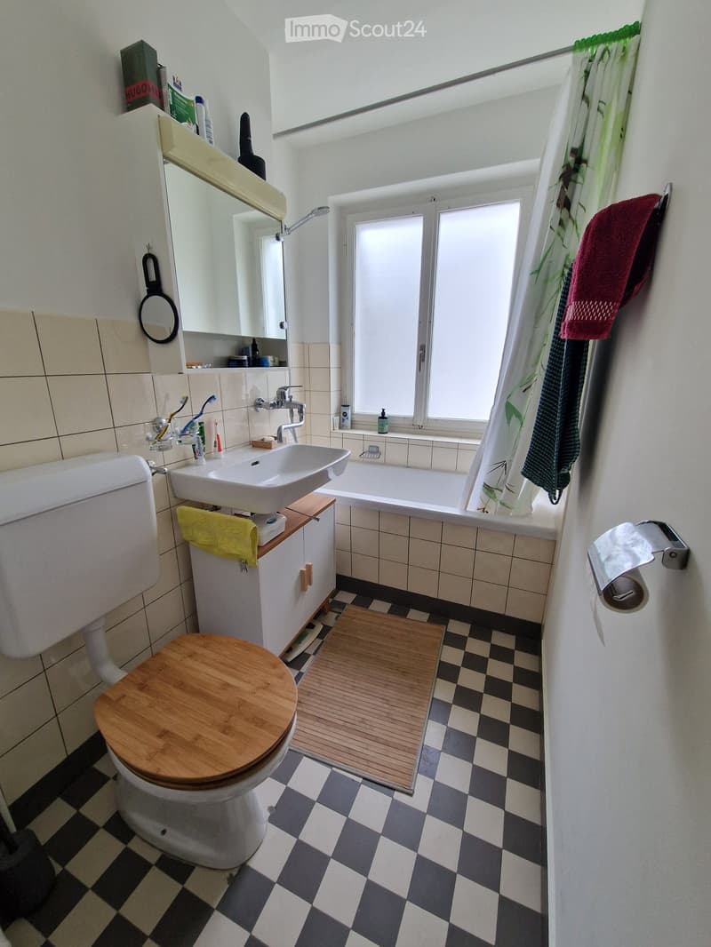 Bathroom with a toilet, sink, bathtub, mirror, and various toiletries. Features a checkered floor and a wooden bath mat.