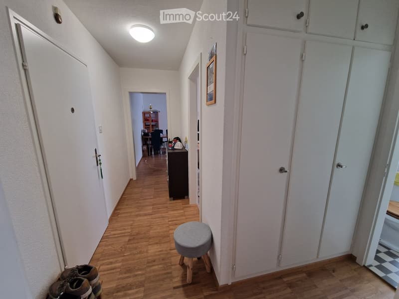 A narrow hallway with wooden flooring, white walls, and several doors. A small grey stool is placed near the center, and a pair of shoes is on the floor to the left. A framed picture hangs on the wall to the right, and a room with a desk and chairs is vis