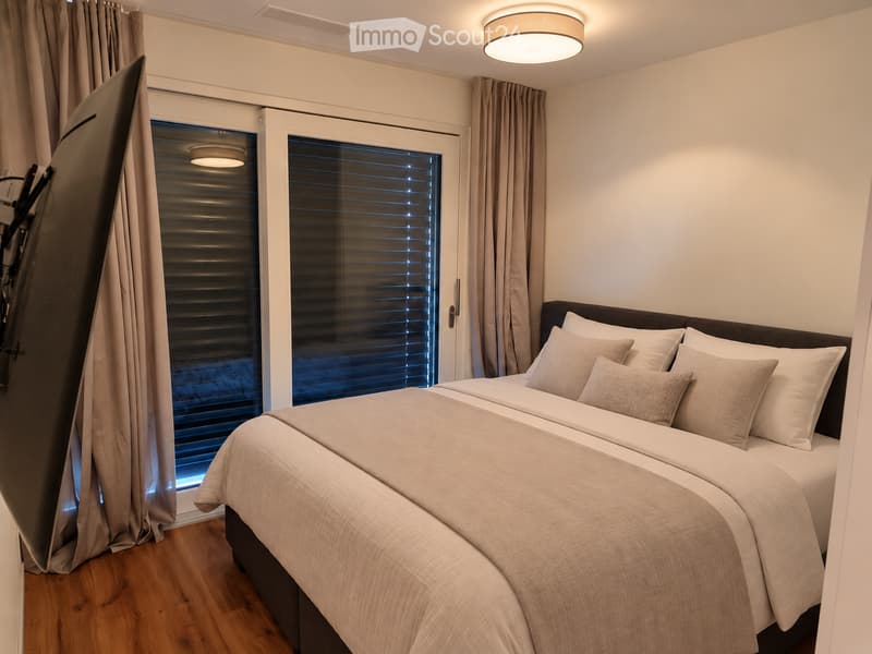 Modern bedroom with a neatly made double bed, multiple pillows, and a large window with blinds and curtains. The room features wooden flooring and a ceiling light fixture.