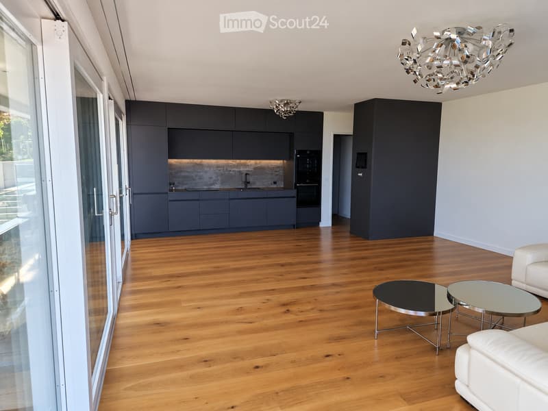 Modern living room with wooden flooring, white leather sofa, two round tables, and an open kitchen area with dark cabinets and stainless steel appliances.