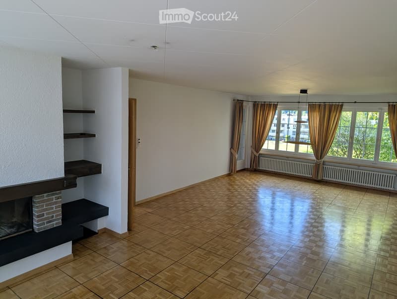 Empty room with wooden flooring, white walls, large windows with curtains, and a brick fireplace with built-in shelves.