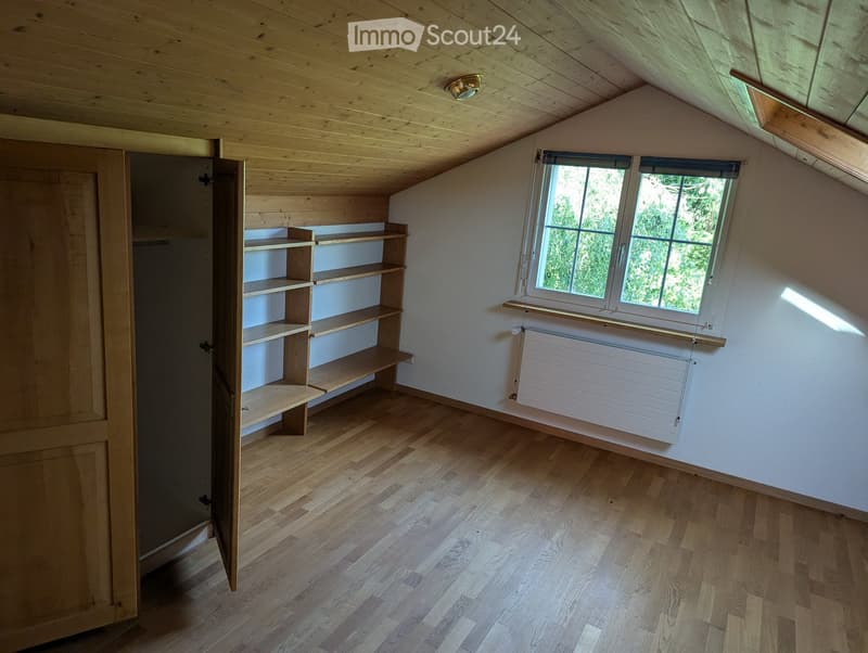 Empty attic room with wooden floors, slanted wooden ceiling, built-in wooden shelves, and a window with a radiator below.