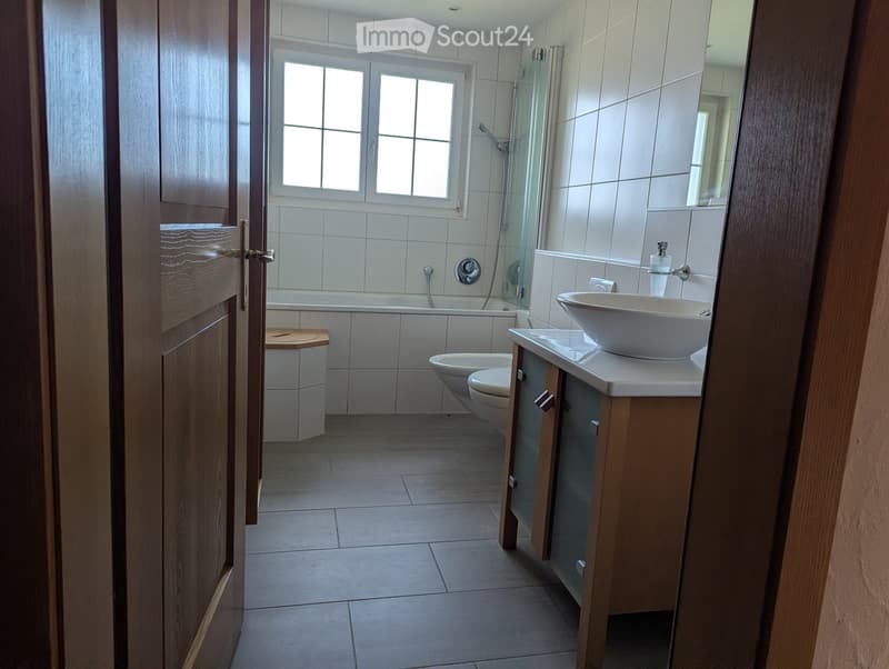 Modern bathroom with white tiles, a bathtub, toilet, sink, and a glass-enclosed shower area. Features a wooden door and a window for natural light.