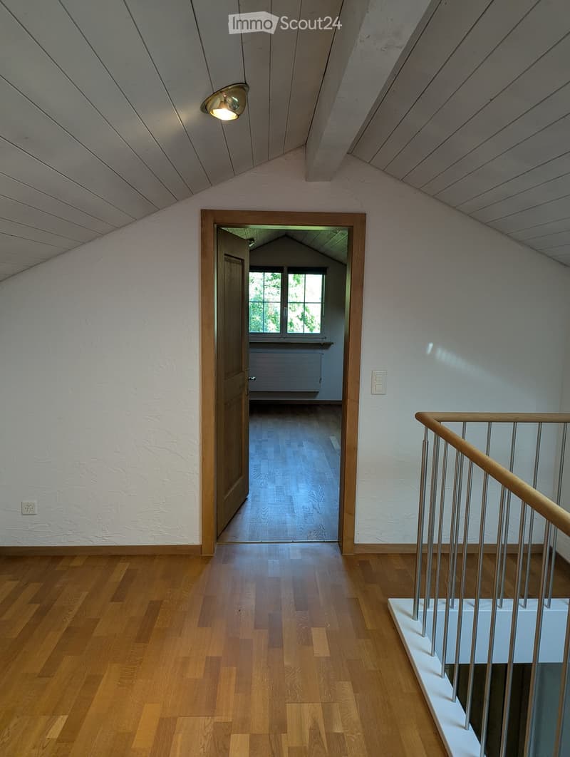 Bright hallway with wooden flooring, white walls, and a staircase leading upwards. A door opens to another room with large windows and wooden flooring.