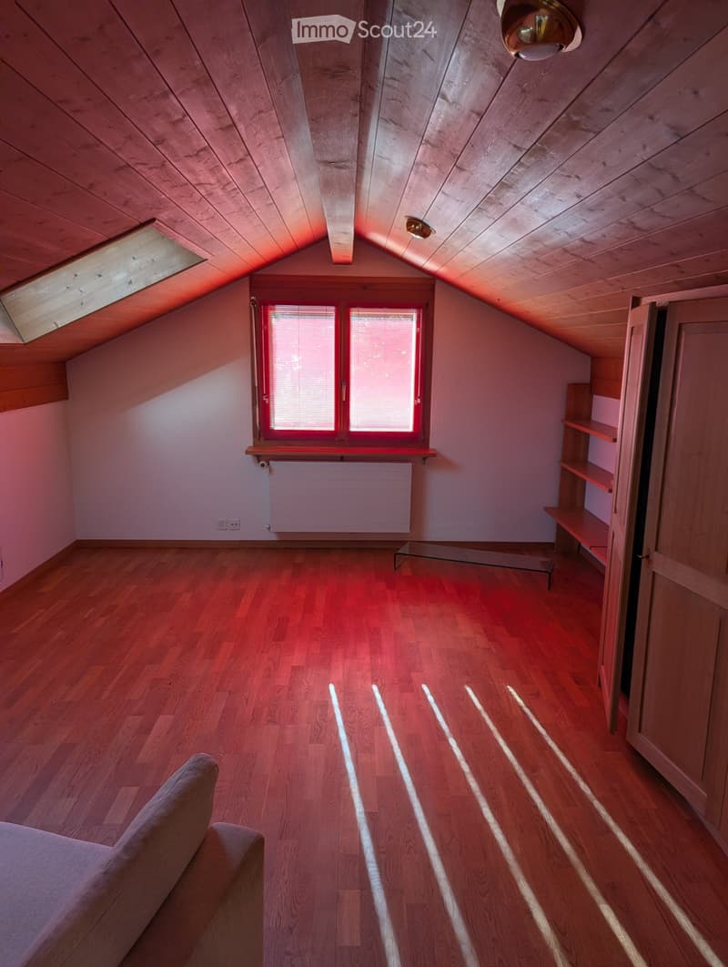 Spacious attic room with wooden floors, slanted wooden ceiling, large windows, built-in shelves, and a sofa.