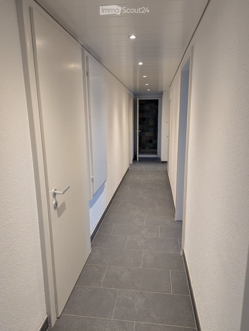 Clean, modern hallway with grey tiled flooring, white walls, and several closed doors. Recessed ceiling lights provide illumination.