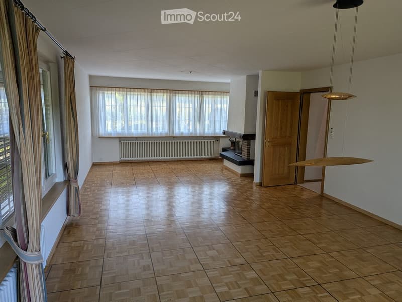 Empty room with wooden flooring, large windows with curtains, radiator, and a hanging light fixture.