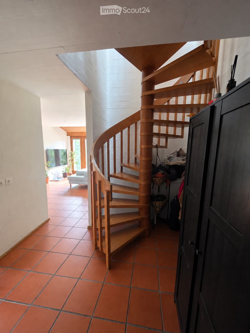 Spiral wooden staircase with open wooden railings, terracotta tiled floor, white brick walls, and a view into a living area with a sofa, TV, and potted plants.
