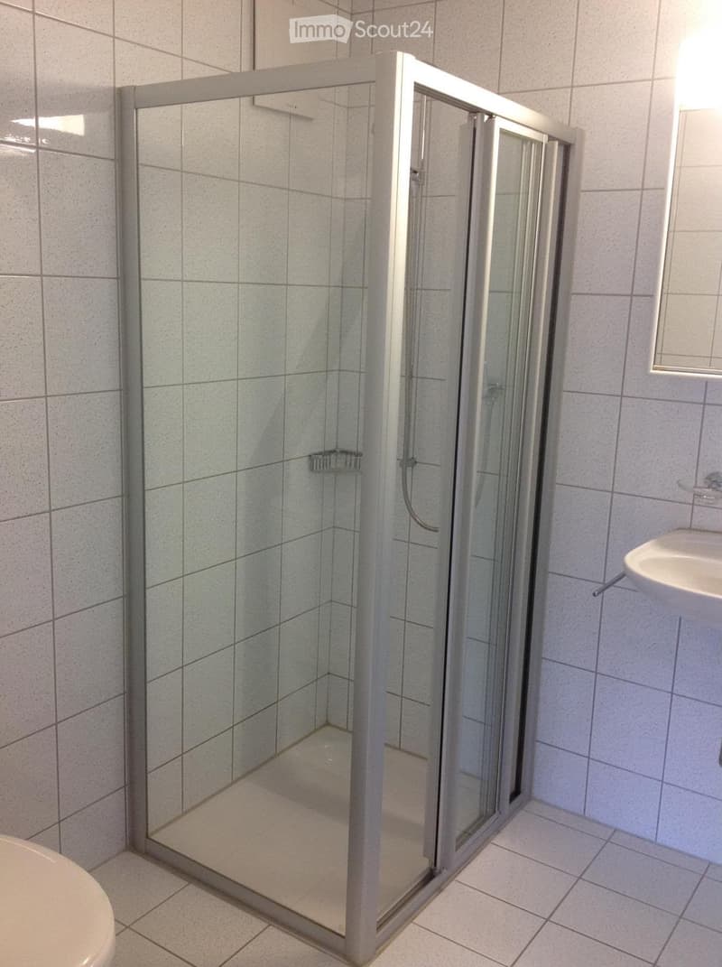 Modern bathroom with a glass-enclosed shower, toilet, and sink. The shower has a built-in shelf and is framed with silver trim. The walls and floor are tiled in a light gray color.