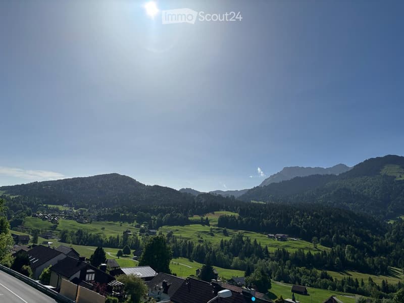 A panoramic view of a lush green valley surrounded by forested mountains, with scattered houses and a winding road in the foreground.