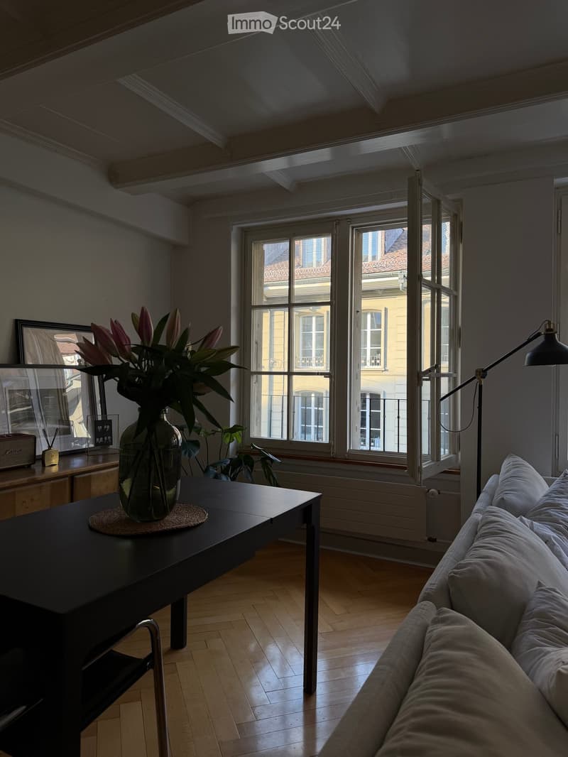 Living room with wooden floor, large windows, sofa, coffee table with vase of flowers, floor lamp, and picture frame on side table.