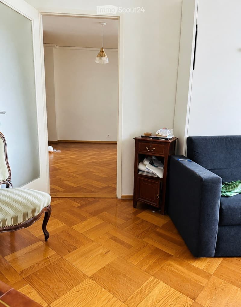 A furnished room with wooden flooring, a sofa, a chair, a small table with a lamp, and a doorway leading to another room.