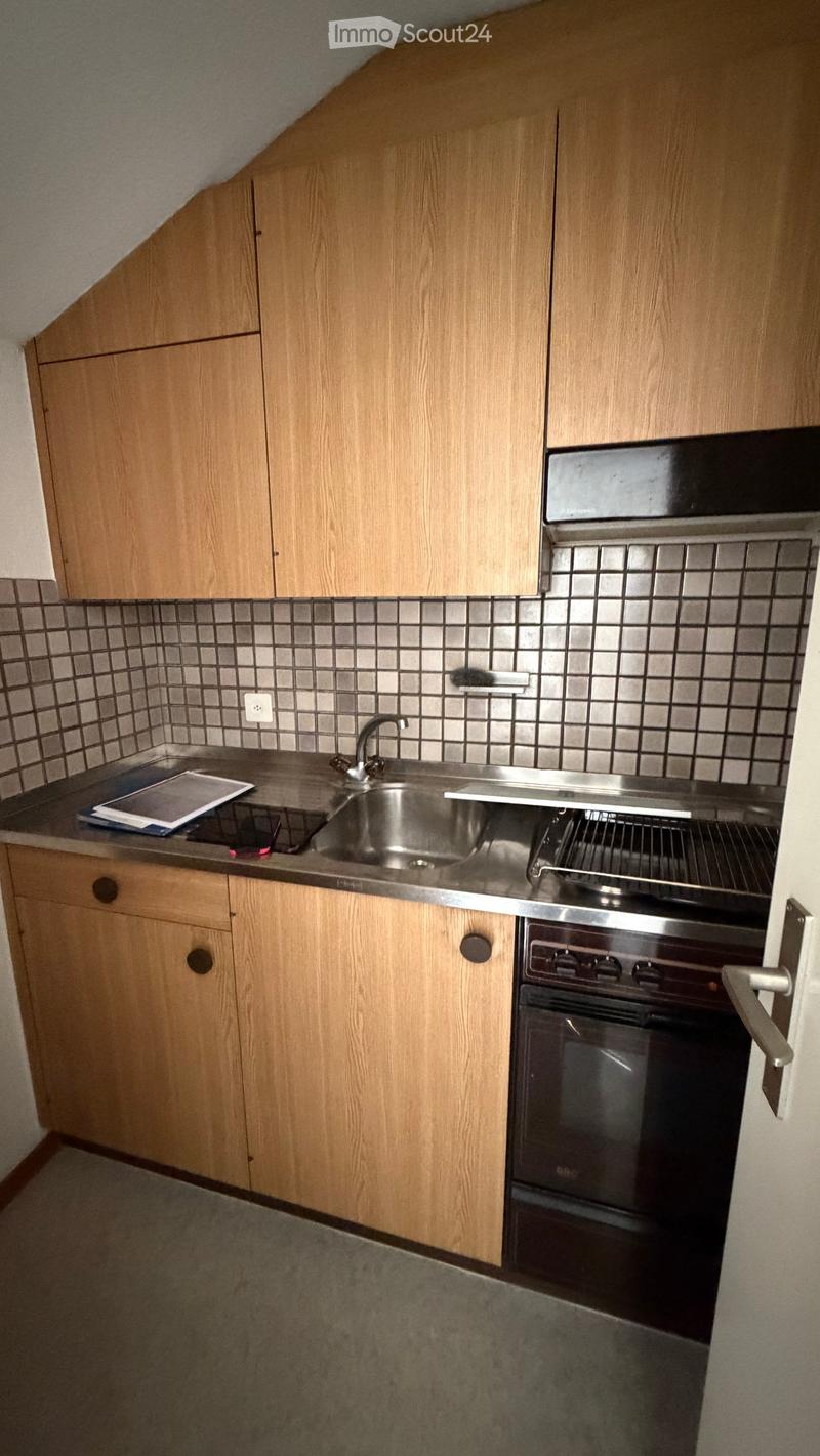 Compact kitchen with wooden cabinets, stainless steel sink, and oven. Countertop has a few items including a tray and papers.