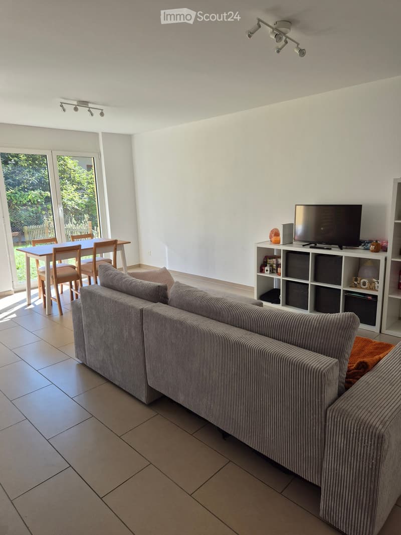Modern living room with a grey sofa, a dining table with chairs, a television on a shelf, and large windows leading to a garden.