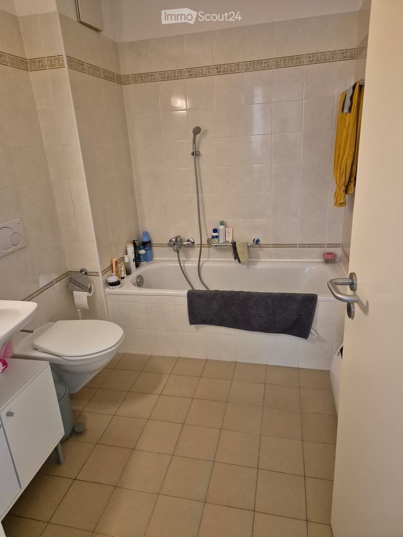 Bathroom with beige tiles, a bathtub with a showerhead, a toilet, a sink, and various toiletries on the edge of the bathtub.