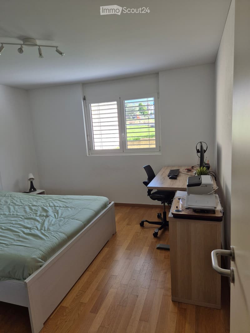 A well-lit bedroom with a bed, desk, chair, and a window with blinds. The room features wooden flooring and modern lighting.