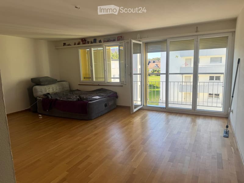 Bright, unfurnished room with wooden flooring, large windows, and a balcony. A sofa with pillows is placed against the wall.