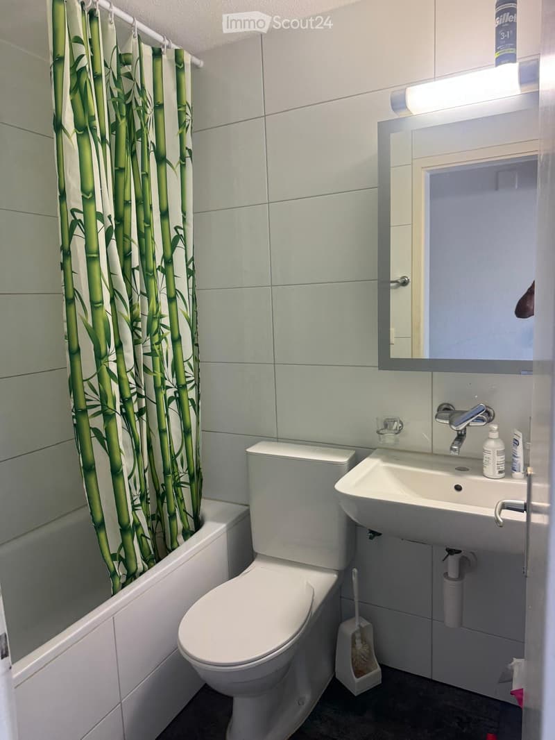 Modern bathroom with white tiles, a toilet, a sink with a mirror, a bathtub with a bamboo-patterned shower curtain, and a light fixture above the mirror.