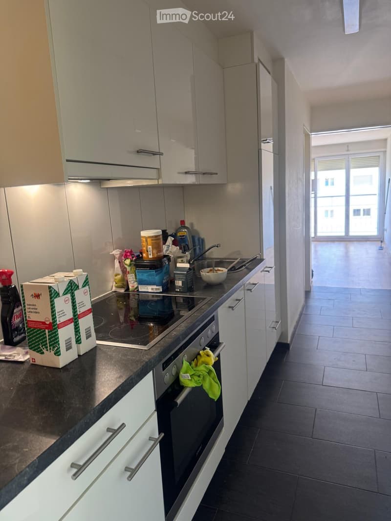 Modern kitchen with white cabinets, black countertops, and stainless steel appliances. Visible items include an oven, fridge, sink, faucet, bowl, and cleaning supplies.