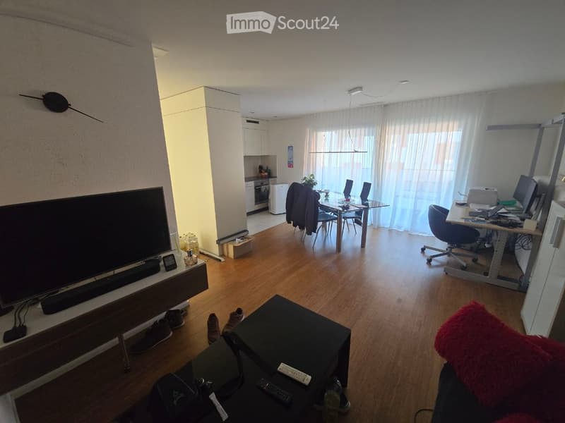 A modern, open-plan living and dining area with a large TV on a stand, a sofa with a red throw pillow, a coffee table, a dining table with chairs, a kitchen area with an oven and refrigerator, and a desk with a computer and chair.