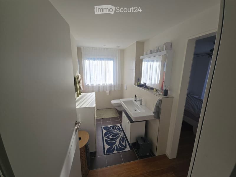 Modern bathroom with a bathtub, toilet, sink, mirror, and cabinet. Light-colored tiles and wooden flooring are visible, along with towels and a decorative rug.