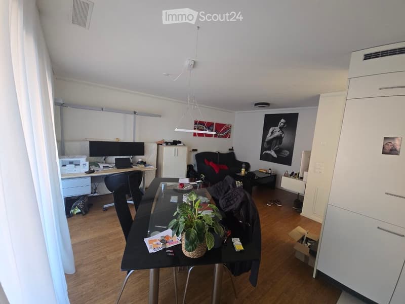 A modern, open-plan living and dining area with a black dining table, chairs, a sofa, a laptop on a desk, and artwork on the walls.