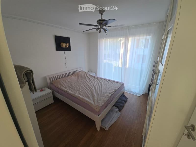 Bedroom with wooden floor, white walls, a bed with a beige duvet, a nightstand, a ceiling fan, and a large window with white curtains.