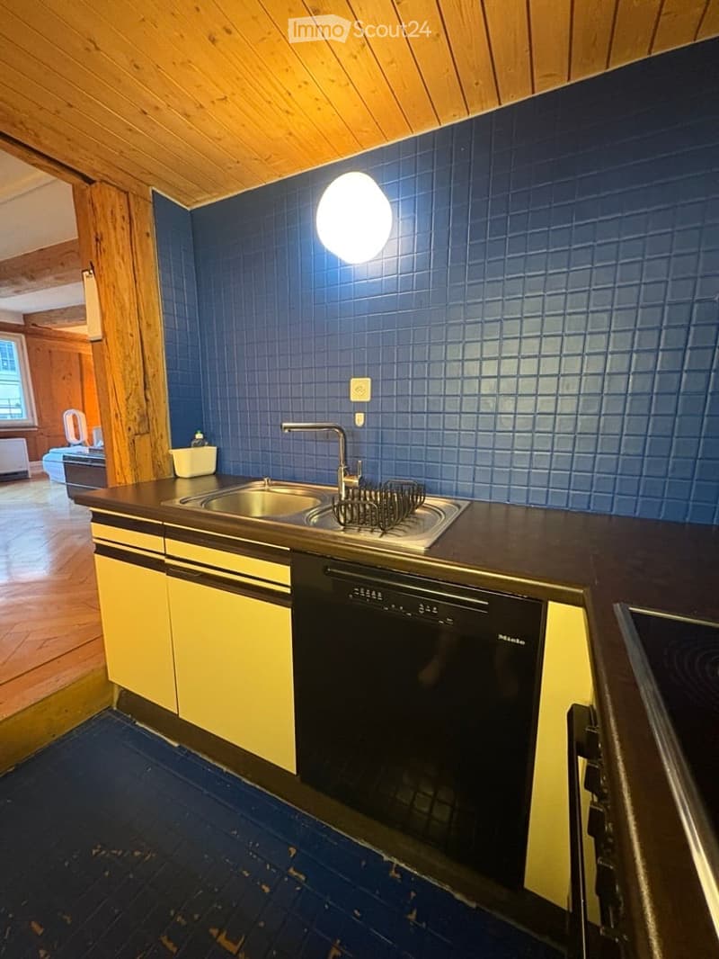 Modern kitchen with blue tiled backsplash, stainless steel sink, and built-in dishwasher. Yellow cabinets and wooden ceiling beams.