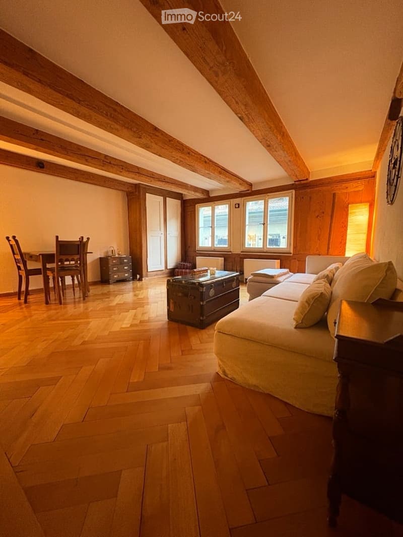 Cozy attic living room with wooden beams, hardwood floor, large windows, sofa, coffee table, dining area with table and chairs, and a small cabinet.