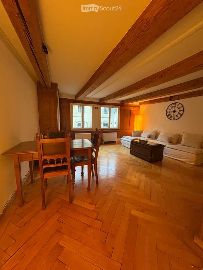 Bright, open room with wooden floors and exposed wooden beams. Features a sofa, a small table with chairs, and a wall-mounted clock.