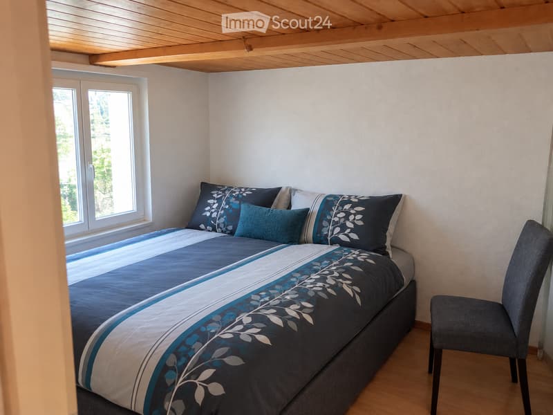 Cozy bedroom with a double bed, blue and white patterned bedding, and a matching chair. The room features light wooden flooring and white walls, with a window offering a view of greenery outside.