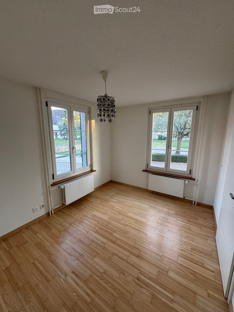 Empty room with wooden flooring, large windows, radiator, and a modern chandelier.
