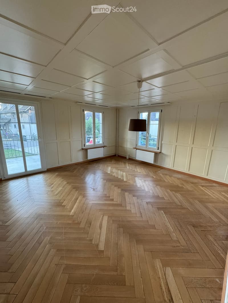 Empty room with wooden herringbone flooring, white walls, large windows, and a hanging light fixture.