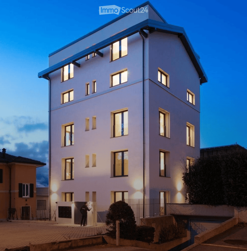 Modern multi-story residential building with illuminated windows and clean lines, featuring a raised ground floor and contemporary design.