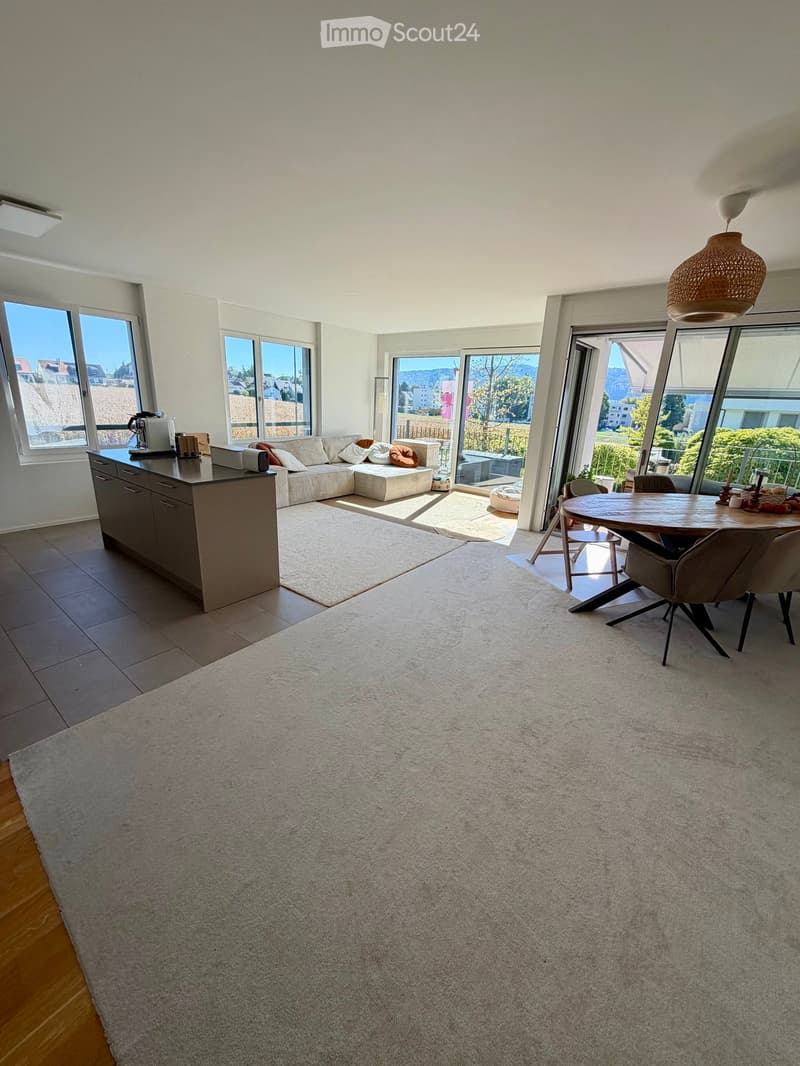 Bright, open living and dining area with large windows offering a scenic view, featuring a sectional sofa, coffee table, kitchen island, dining table, and pendant light.