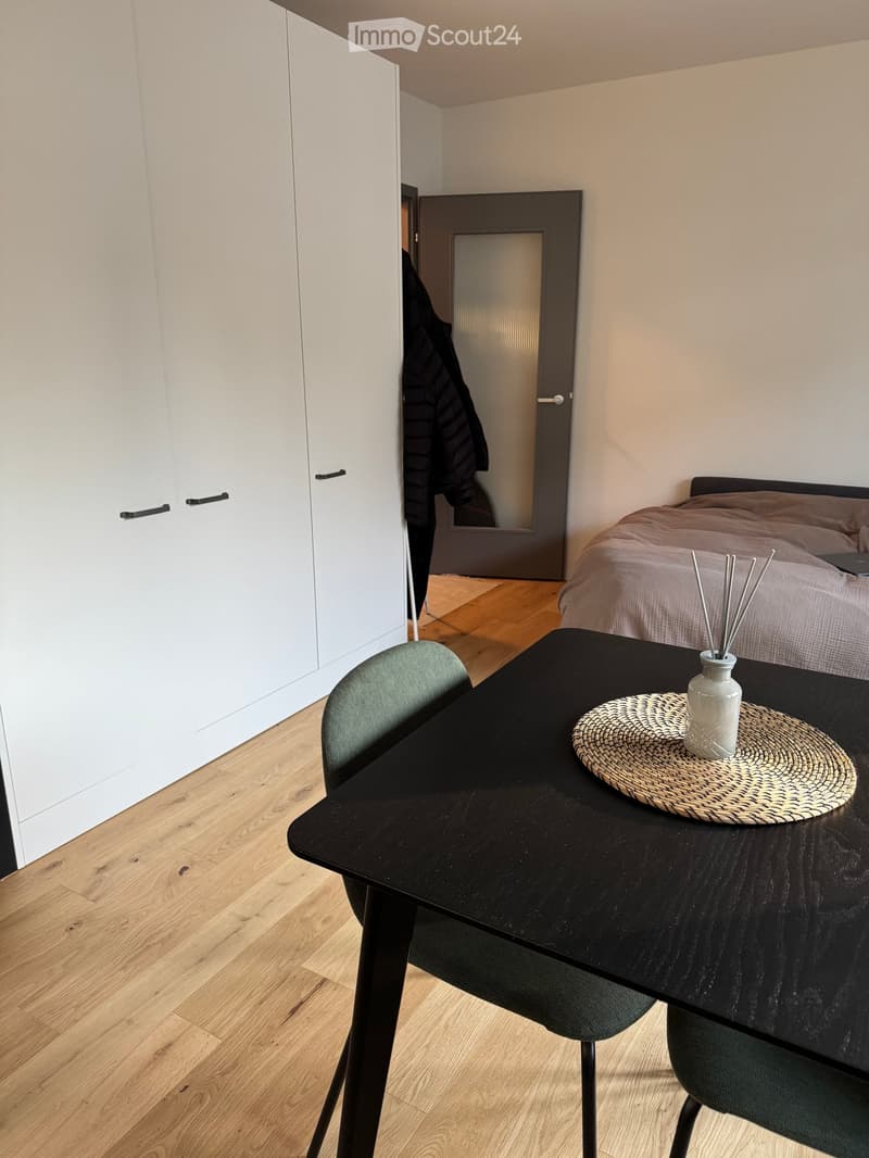 Modern bedroom with a bed, a black table, a chair, a vase on the table, and a placemat. The room features light wooden flooring and white built-in wardrobes.