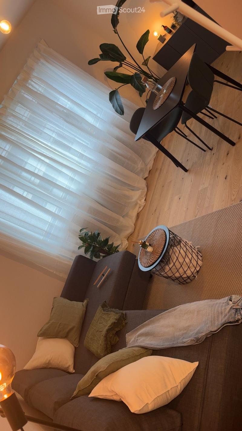 A cozy living room with a grey sectional sofa, multiple pillows, a small black table, a wicker side table, and a potted plant near the window with sheer curtains.