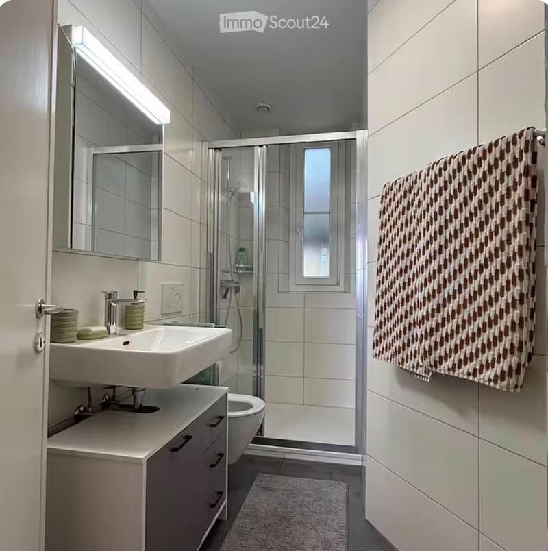 Modern bathroom with white tiles, a double sink, a large mirror, a shower, and a towel hanging on the wall.