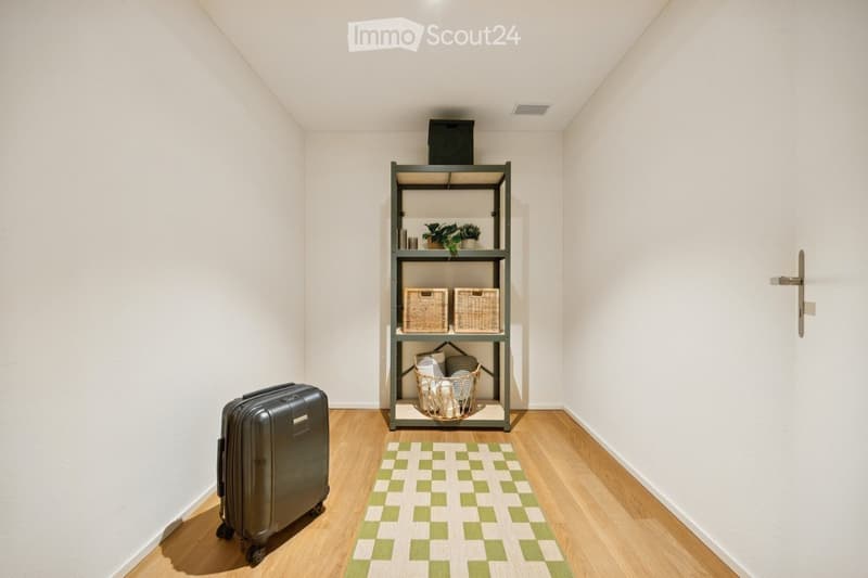 A small, neatly arranged room with light-colored walls and wooden flooring. A metal shelf holds baskets, plants, and other small items. A patterned rug lies on the floor, and a large suitcase is positioned near the wall.